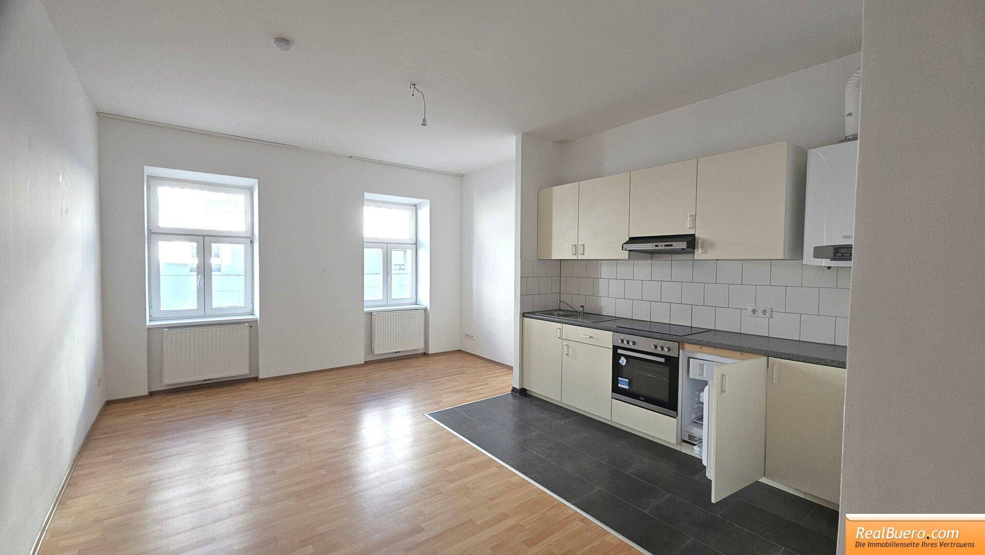 Heller Wohnbereich mit zwei Fenstern, Laminatboden und integrierter Küchenzeile.