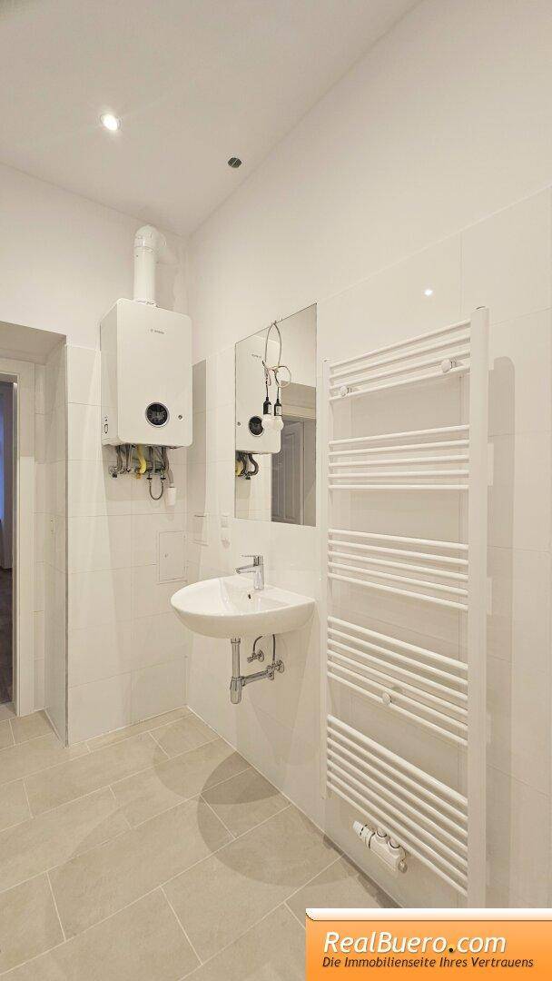 Badezimmer mit Waschbecken, Spiegel und Heizungsanlage an der Wand, sowie einem Handtuchheizkörper.