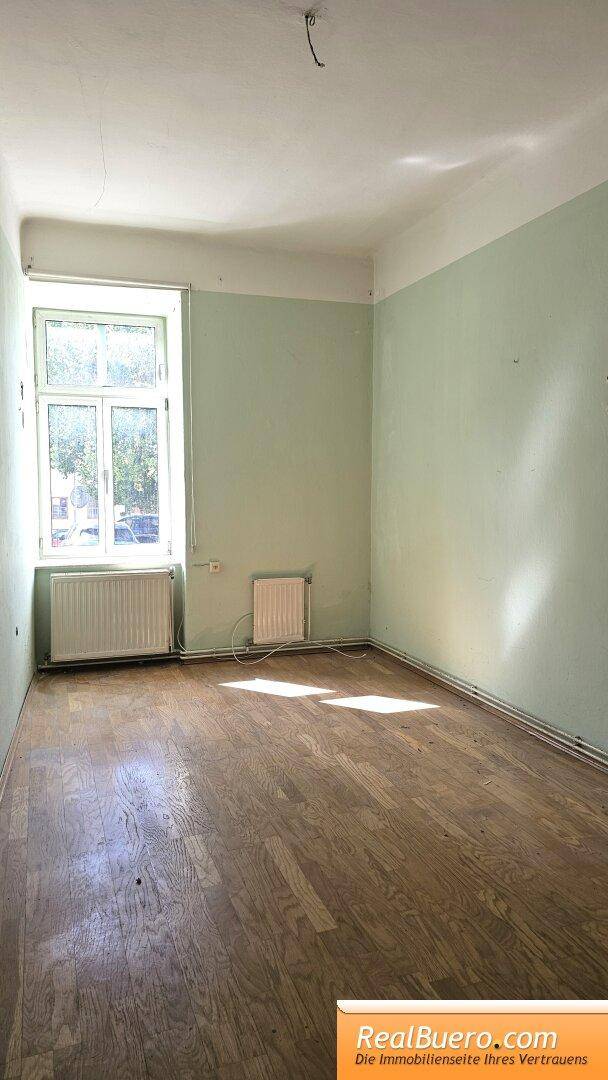 Helles Zimmer mit Holzplankenboden, hellgrünen Wänden und einem Fenster mit Heizkörper.