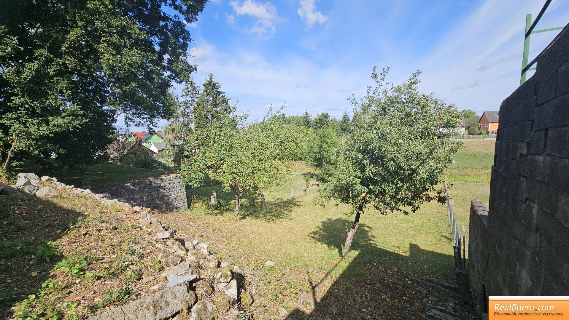 Gartenbereich mit Obstbäumen, trockenem Gras und einer Steinmauer, mit Blick auf Häuser.