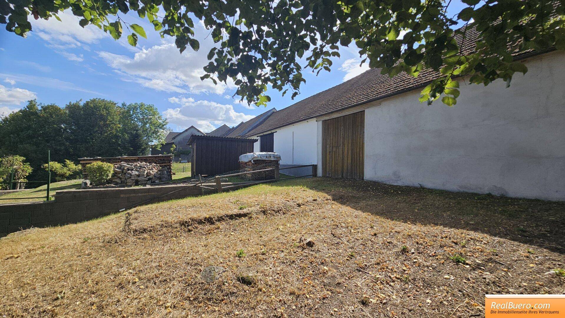 Außenansicht eines Gebäudes mit großer Holztür, umgeben von trockenem Gras und Bäumen.