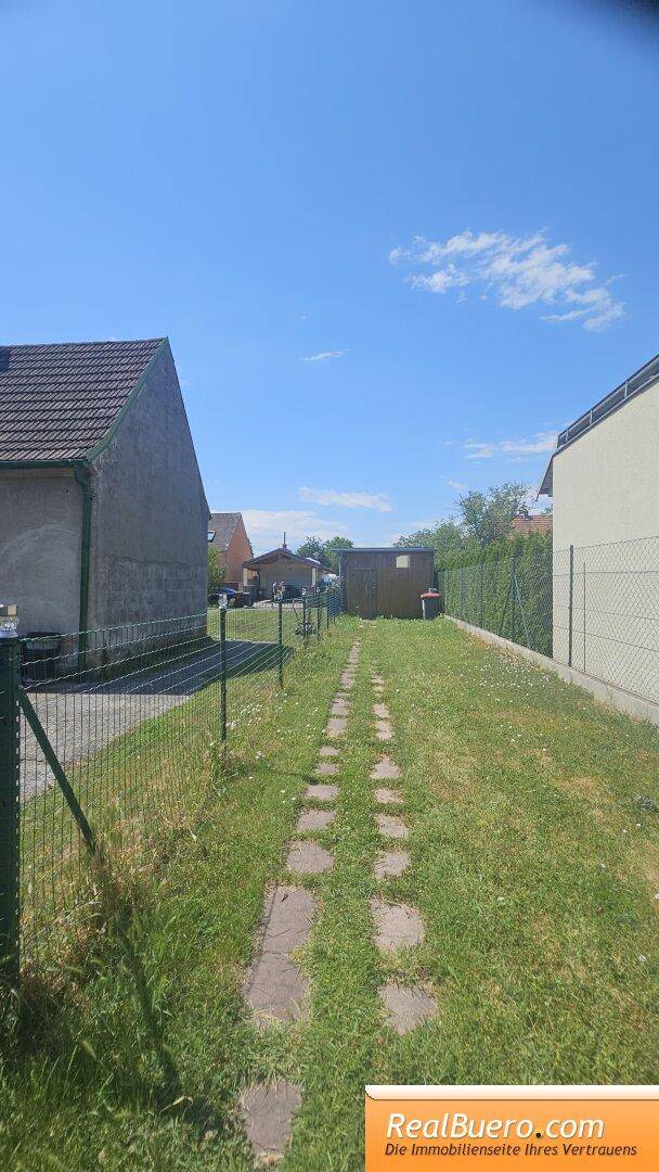 Langer, schmaler Gartenbereich mit einem Steinweg, beidseitig eingezäunt und einem Schuppen im Hintergrund.