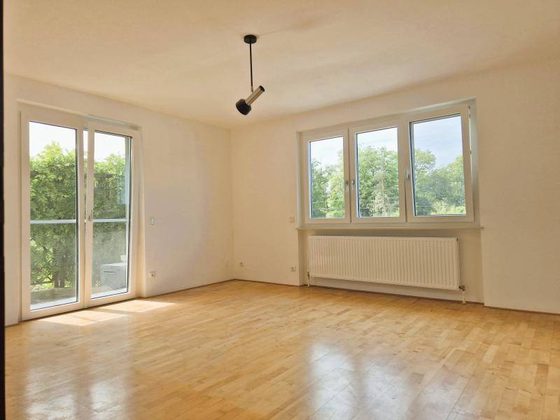 Leerer Raum mit Holzboden, großen Fenstern und Zugang zu einem Balkon.