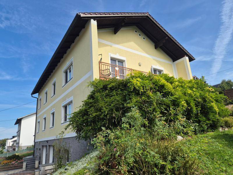 Seitenansicht des Hauses mit Balkon, umgeben von üppigem Grün und Sträuchern unter blauem Himmel.