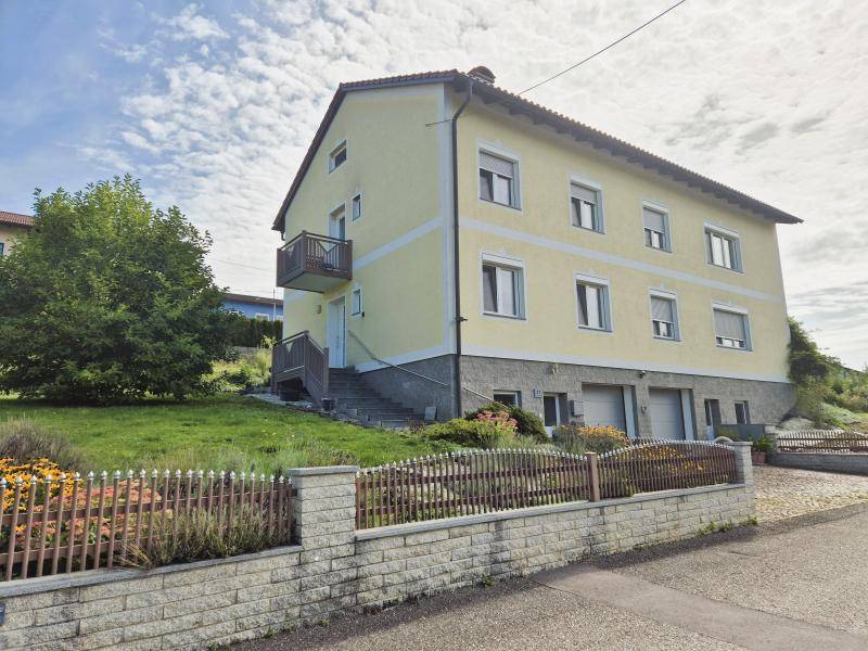 Gelbes Wohnhaus mit Außentreppe, Garagen und einem kleinen Vorgarten unter bewölktem Himmel.