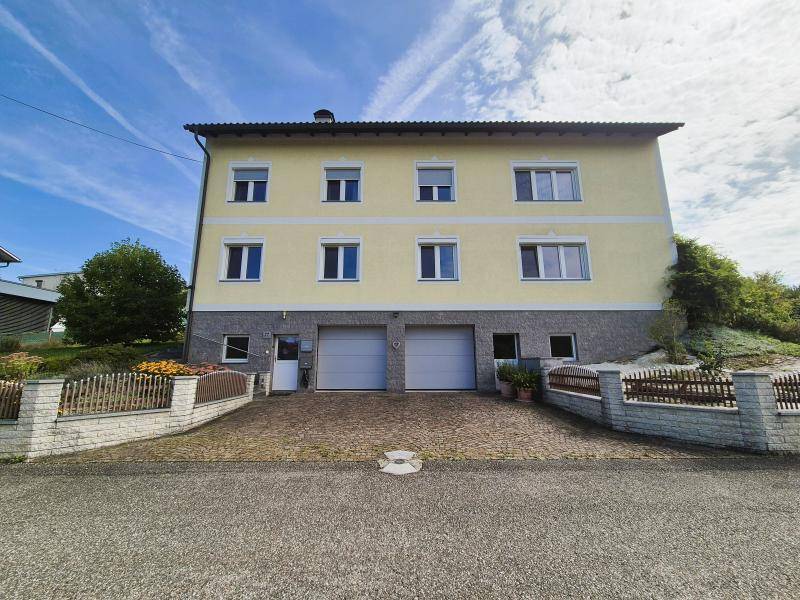 Vorderansicht eines gelben Hauses mit zwei Garagen und gepflasterter Auffahrt unter blauem Himmel.