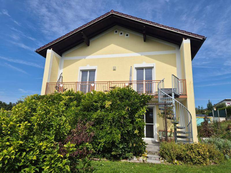 Seitenansicht des Hauses mit Balkon, teilweise von hohen Büschen verdeckt, unter blauem Himmel.