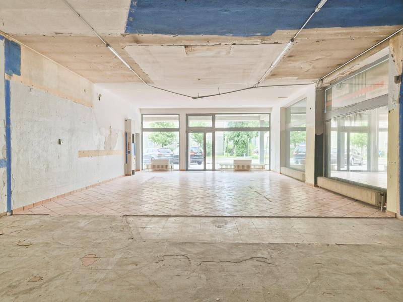 Großer, leerstehender Ladenraum mit Fliesenboden, großen Schaufenstern und sichtbaren Deckeninstallationen.