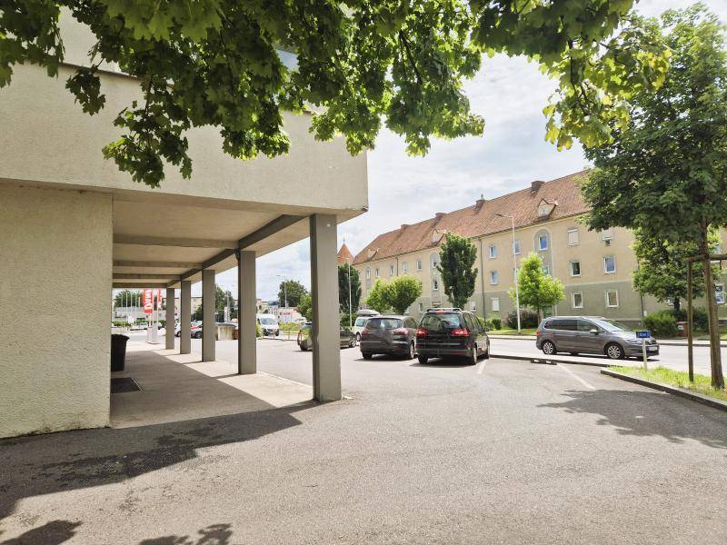 Seitenansicht des Gebäudes mit überdachtem Gehweg und parkenden Autos, umgeben von Bäumen und Wohngebäuden.
