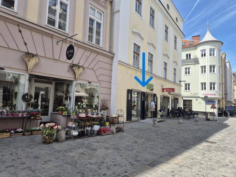 Historisches Gebäude mit Ladenlokal im Erdgeschoss, umgeben von anderen Gebäuden und einer gepflasterten Straße.