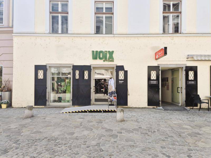 Frontansicht des Ladenlokals mit Schaufenstern, Eingangstür und einer Rampe auf der gepflasterten Straße.