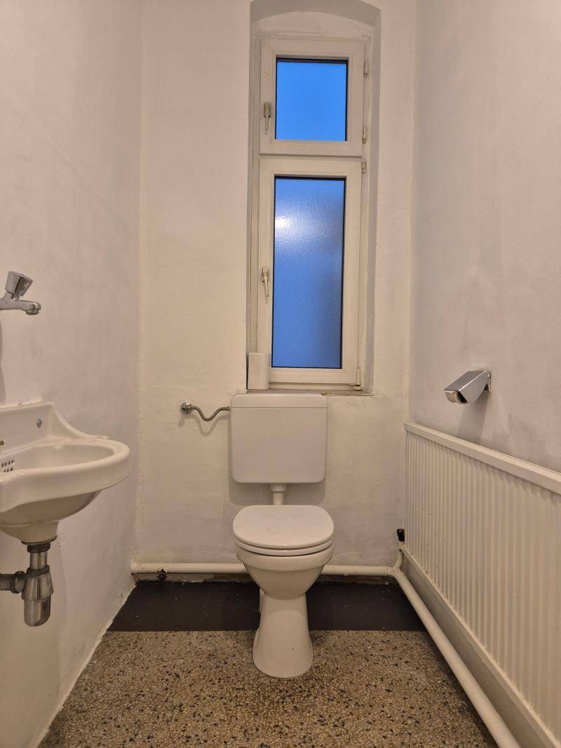 Einfache Toilette mit Fenster, Waschbecken und Heizkörper, funktional und hell.