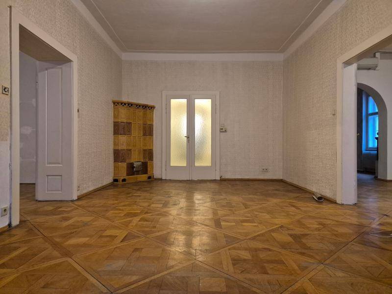 Leerer Wohnraum mit einem traditionellen Kachelofen und eleganten Doppeltüren, die den Zugang zu anderen Räumen ermöglichen.