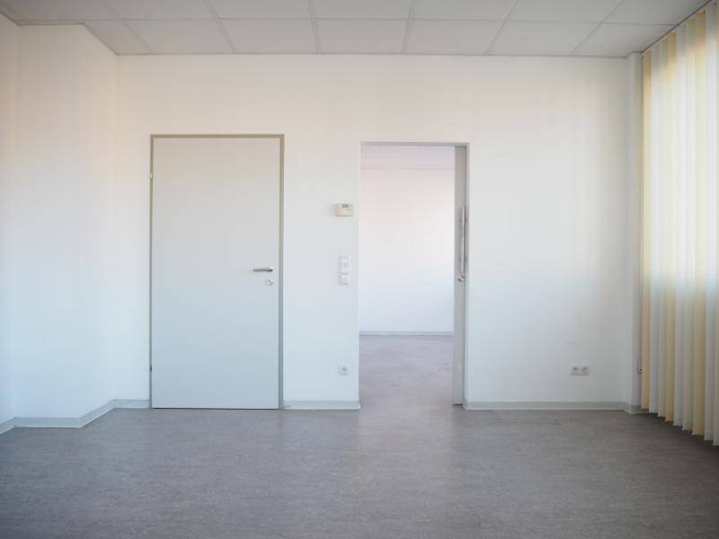 Leerer Büroraum mit weiß gestrichenen Wänden, grauem Boden und zwei Türen zu angrenzenden Bereichen.
