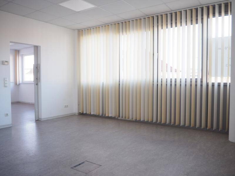 Geräumiger Büroraum mit hellem Vinylboden, vertikalen Jalousien und Zugang zu einem weiteren Zimmer.