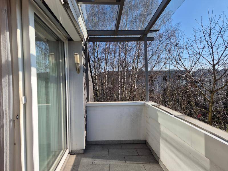 Heller Balkon mit Fliesenboden und Glasüberdachung, umgeben von Bäumen.