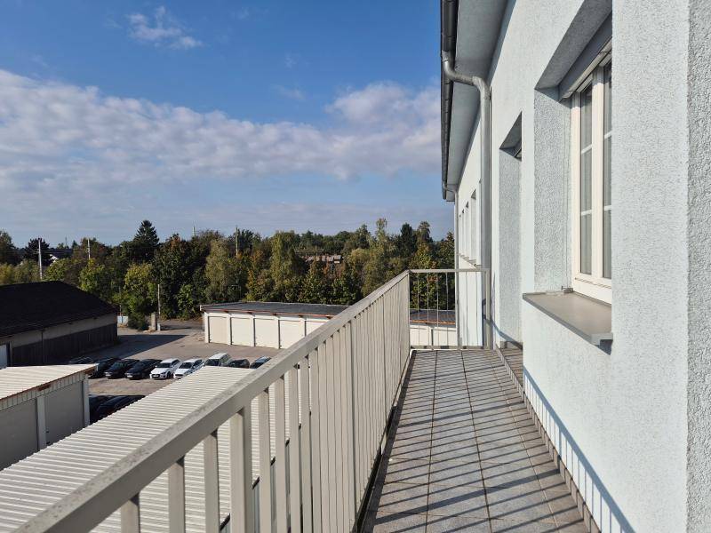 Langer Balkon mit Metallgeländer und Blick auf Bäume und Parkplätze unter blauem Himmel.