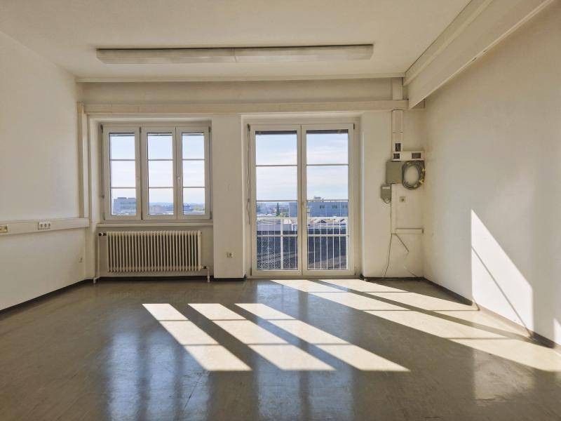 Großer, heller Büroraum mit zwei Fenstern und Balkonzugang, der viel natürliches Licht bietet.