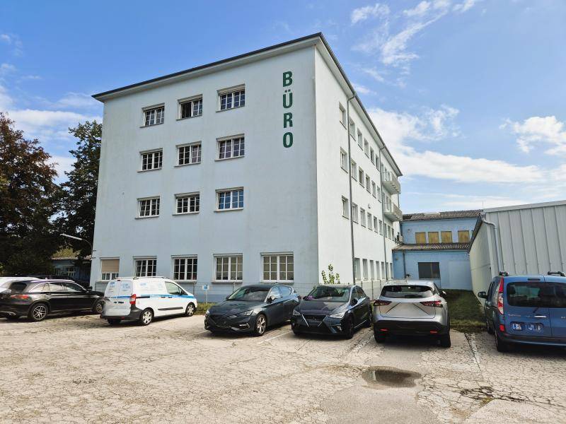 Seitenansicht eines modernen Bürogebäudes mit Parkplätzen und Bäumen im Hintergrund.