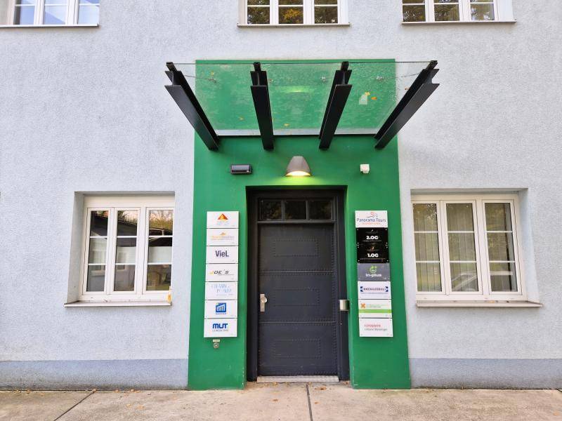 Eingangsbereich eines Gebäudes mit grüner Fassade, einer dunklen Tür und mehreren Firmenschildern.