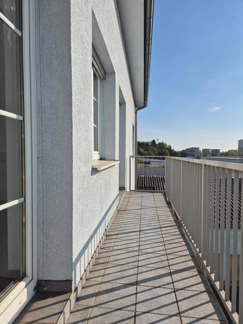 Schmaler Balkon mit Fliesenboden und Metallgeländer entlang der Gebäudefassade bei Sonnenschein.
