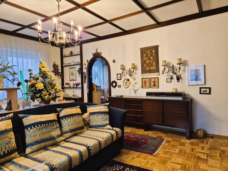 Wohnzimmer mit bequemer Couch, Sideboard und dekorativen Wandelementen unter Holzbalkendecke.
