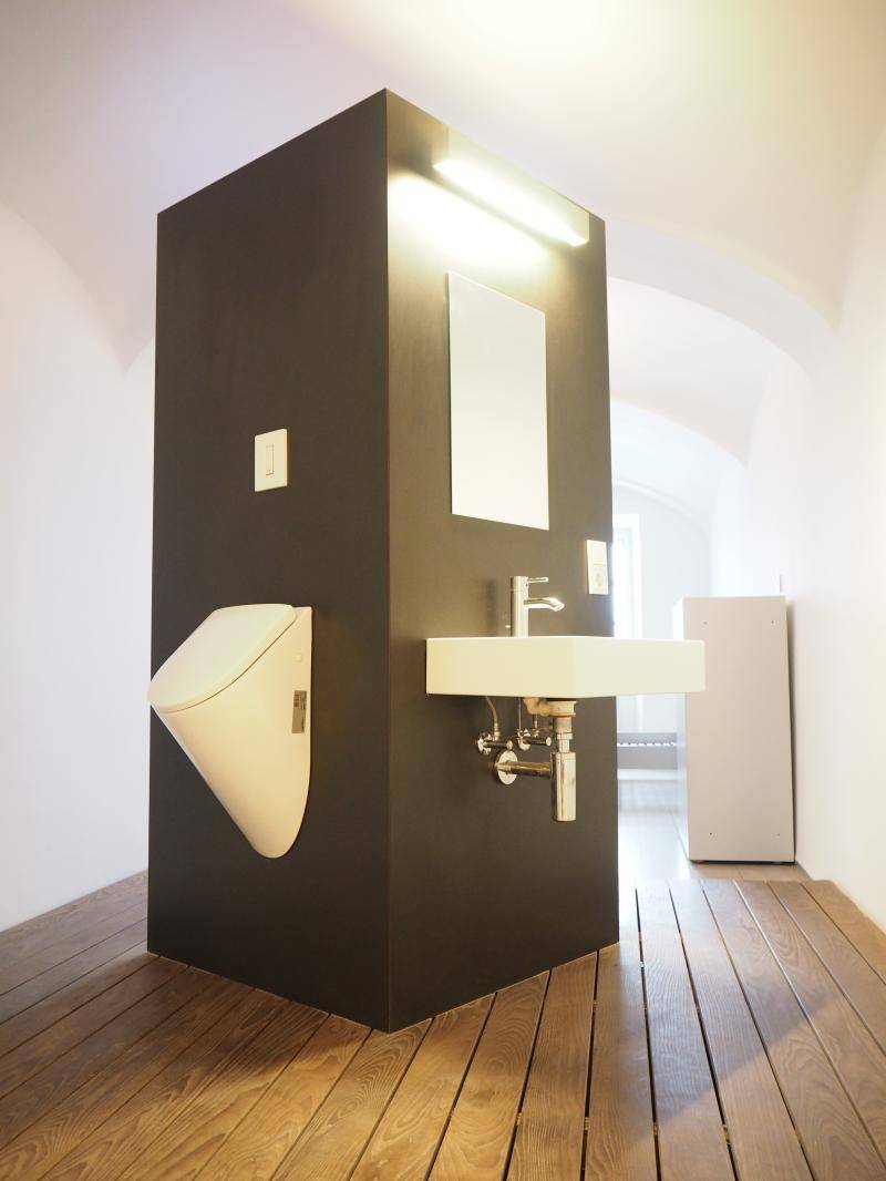 Modernes WC mit Urinal, Waschbecken und dunkler Trennwand auf Holzboden.