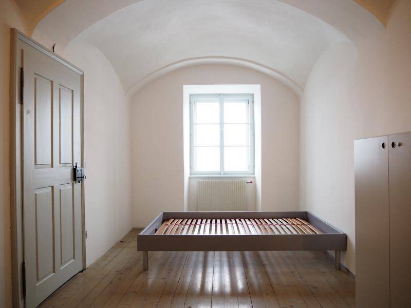 Schlafzimmer mit Gewölbedecke, Holzboden, Fenster und einfachem Bettrahmen.