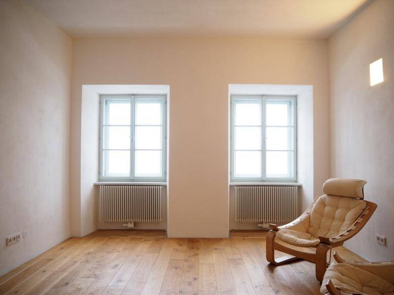 Helles Wohnzimmer mit Holzboden, zwei Fenstern und einem bequemen Sessel, schlicht eingerichtet.