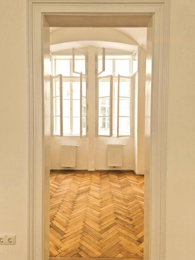 Blick durch einen Türrahmen in einen hellen Raum mit Fischgrätparkett und großen Fenstern.