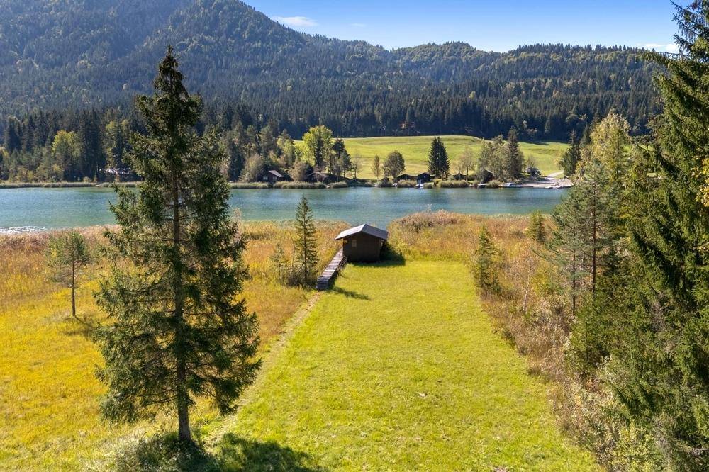 Luftaufnahme eines kleinen Holzhauses mit Steg am Seeufer, umgeben von grünen Wiesen und Wäldern.