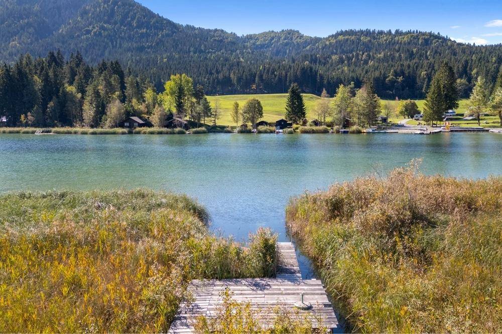 Panoramablick auf den ruhigen Bergsee mit bewaldeten Ufern und majestätischen Bergen im Hintergrund.
