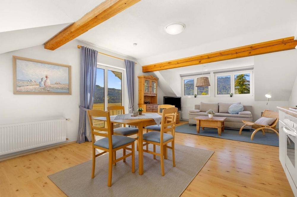 Heller Wohn-Essbereich mit Holzboden, sichtbaren Deckenbalken und Zugang zum Balkon mit Bergblick.
