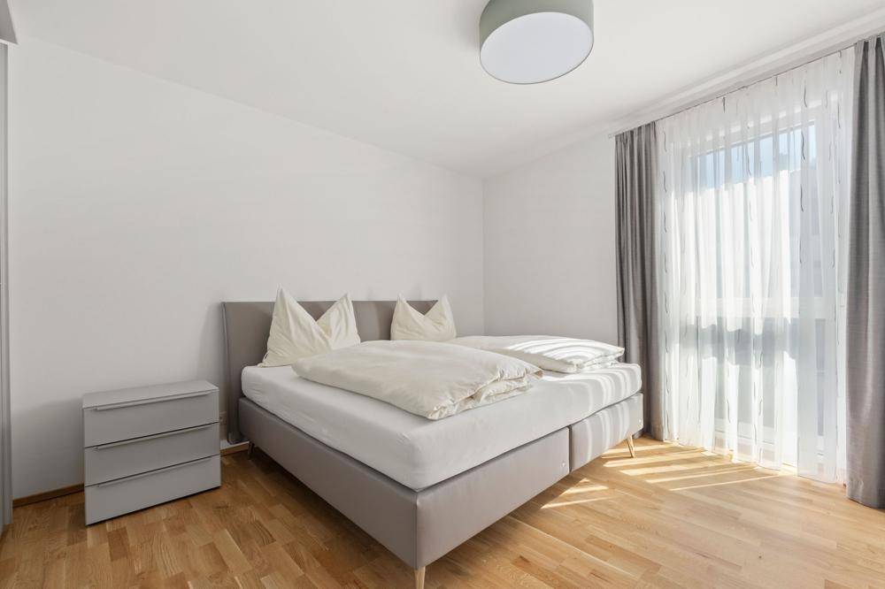 Helles Schlafzimmer mit Doppelbett, Holzboden und großem Fenster mit Vorhängen.