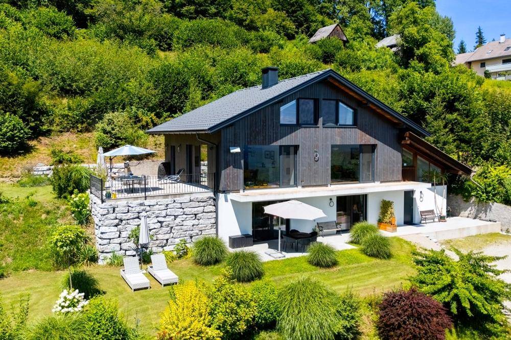 Modernes Haus mit dunkler Holzfassade, großen Fenstern und mehreren Terrassenbereichen im Grünen.