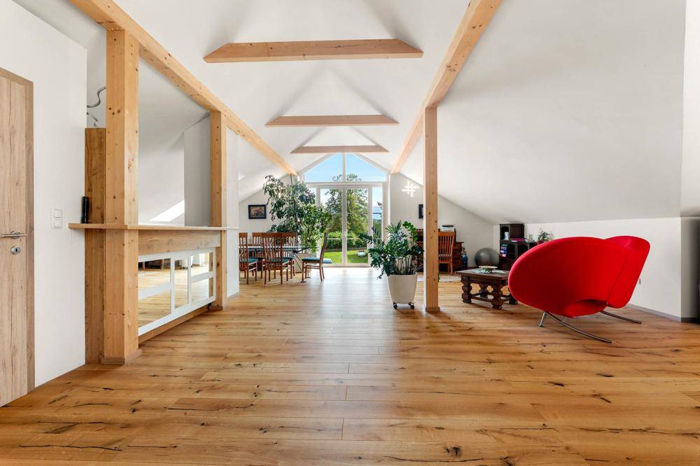 Geräumiger Dachboden mit sichtbaren Holzbalken, hohen Decken und großen Fenstern mit Blick ins Grüne.
