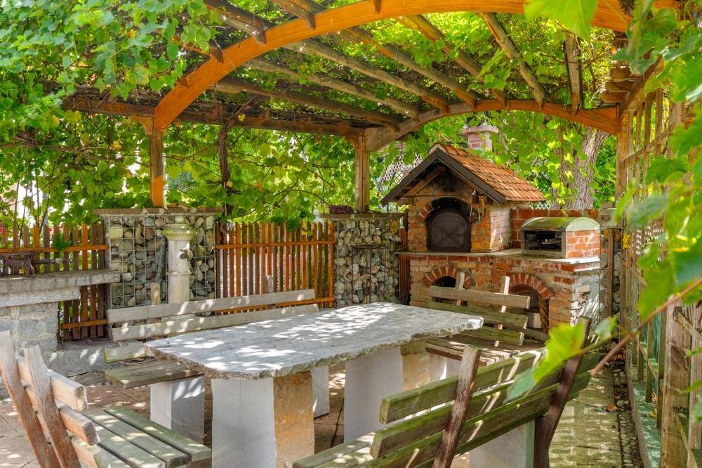 Rustikaler Außenbereich mit Pergola, Steintisch und gemauertem Pizzaofen, umgeben von üppigem Grün.