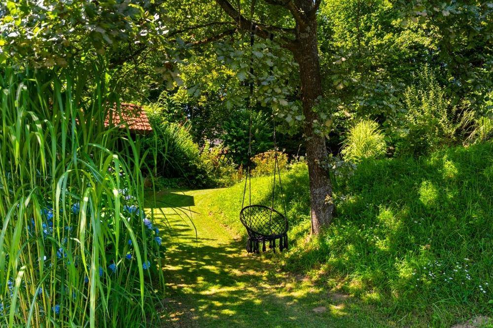 Idyllischer Gartenbereich mit Schaukelstuhl unter einem Baum, umgeben von üppigem Grün.
