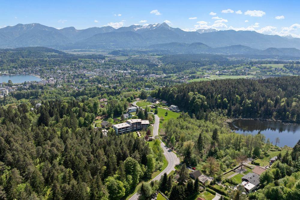 Weitläufige Luftaufnahme einer Siedlung in einer grünen Landschaft mit Seen und majestätischen Bergen im Hintergrund.