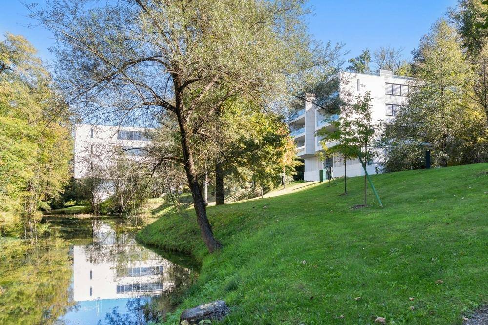Moderne Wohngebäude in grüner Umgebung mit Wasserlauf, umgeben von Bäumen und Rasenflächen.