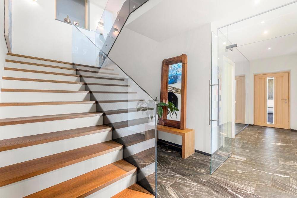Moderner Eingangsbereich mit Holztreppe, Glasgeländer und dunklem Steinboden.
