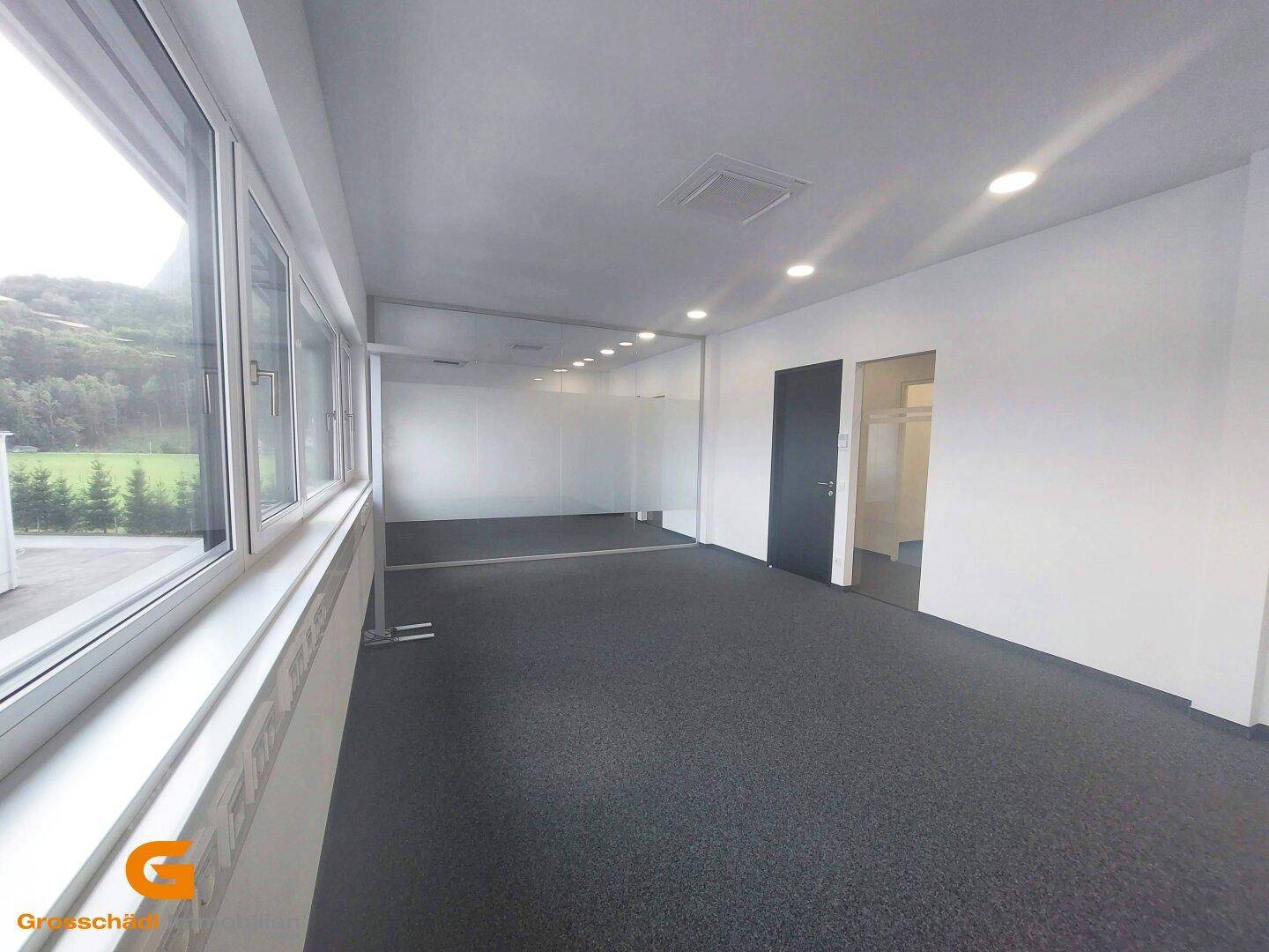 Helles Büro mit langer Fensterfront, Teppichboden und modernen Glaswänden.