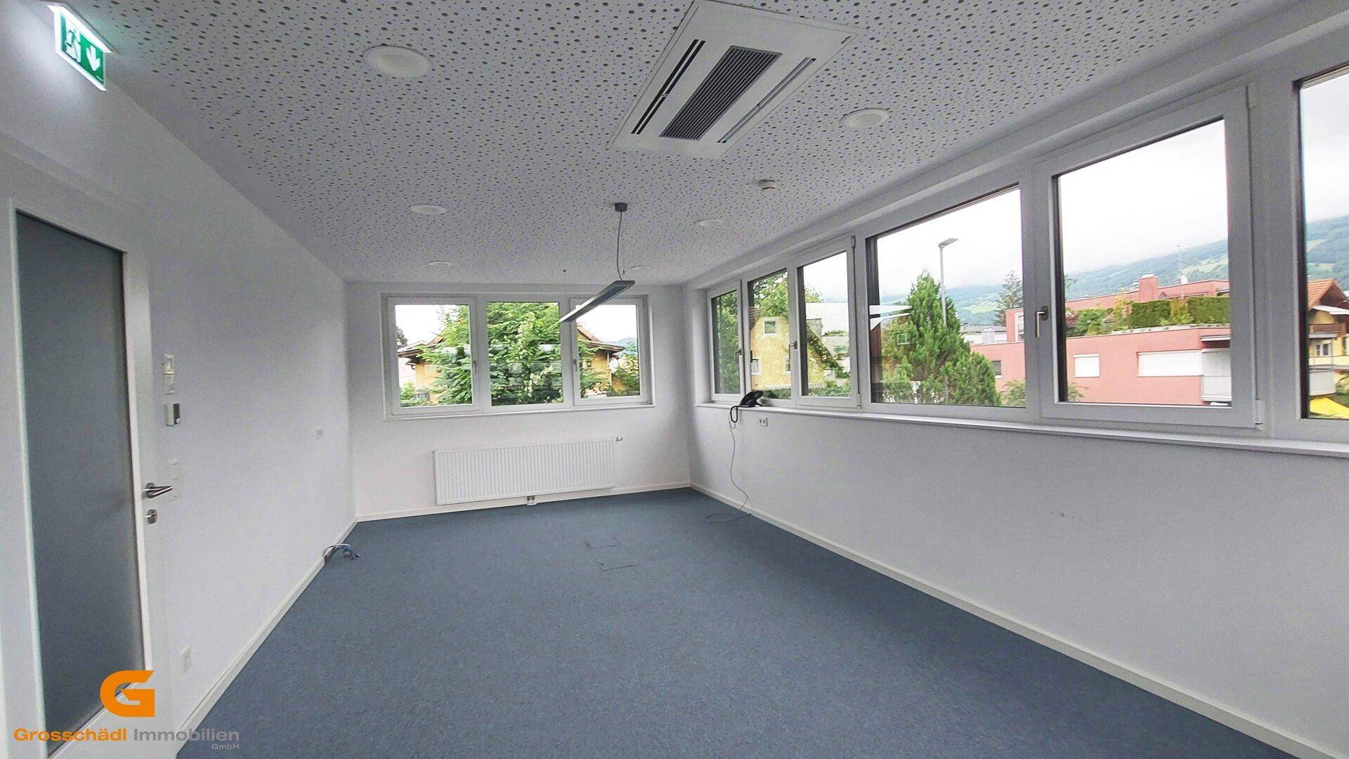 Heller Büroraum mit großen Fenstern, blauem Teppichboden und Klimaanlage an der Decke.