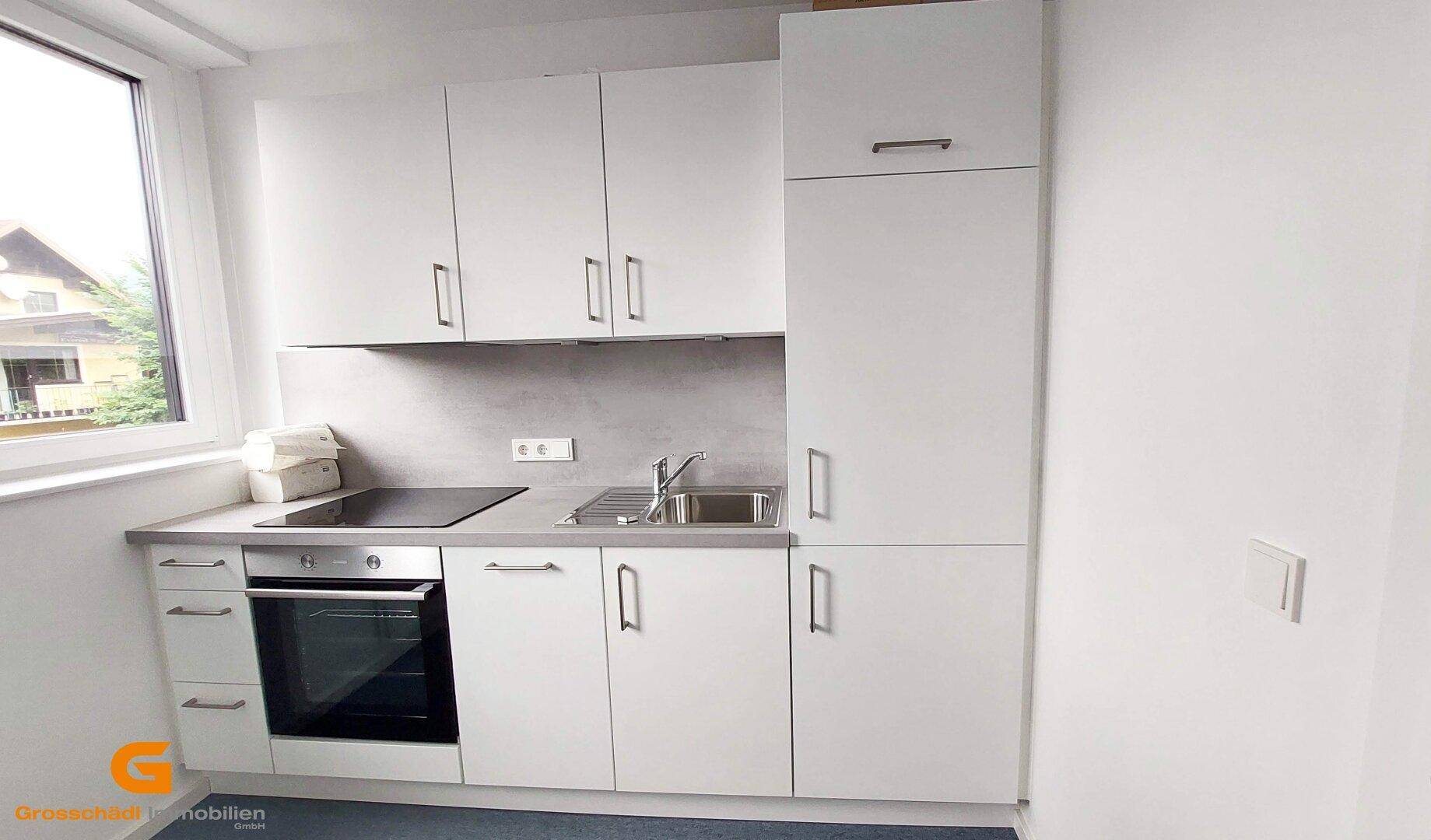 Moderne Küchenzeile mit weißen Schränken, Induktionskochfeld, Backofen und Spüle, neben einem Fenster.