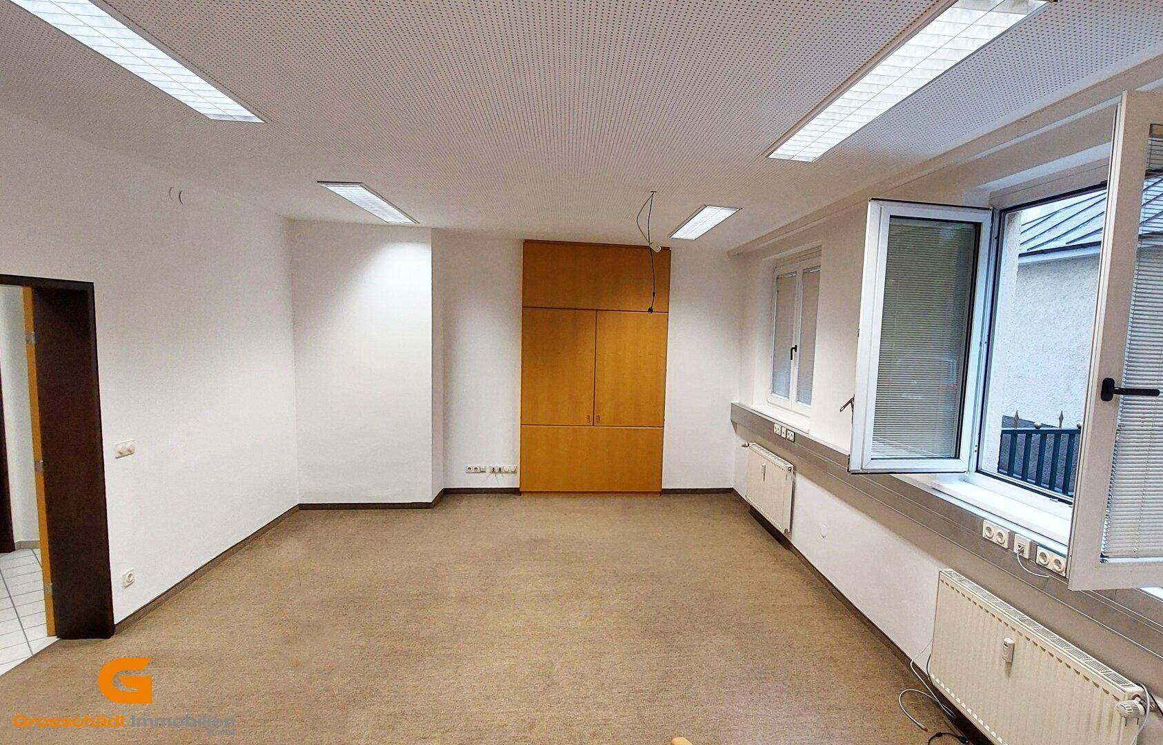 Heller Büroraum mit Teppichboden, Einbauschrank und geöffneten Fenstern für Frischluft.