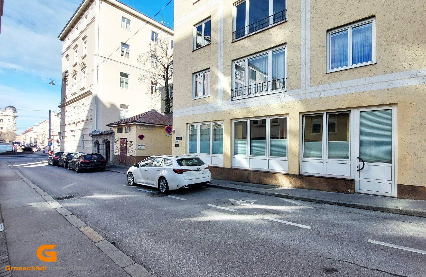 Gebäudefassade mit großen Fenstern und parkenden Autos auf der Straße in einer städtischen Umgebung.