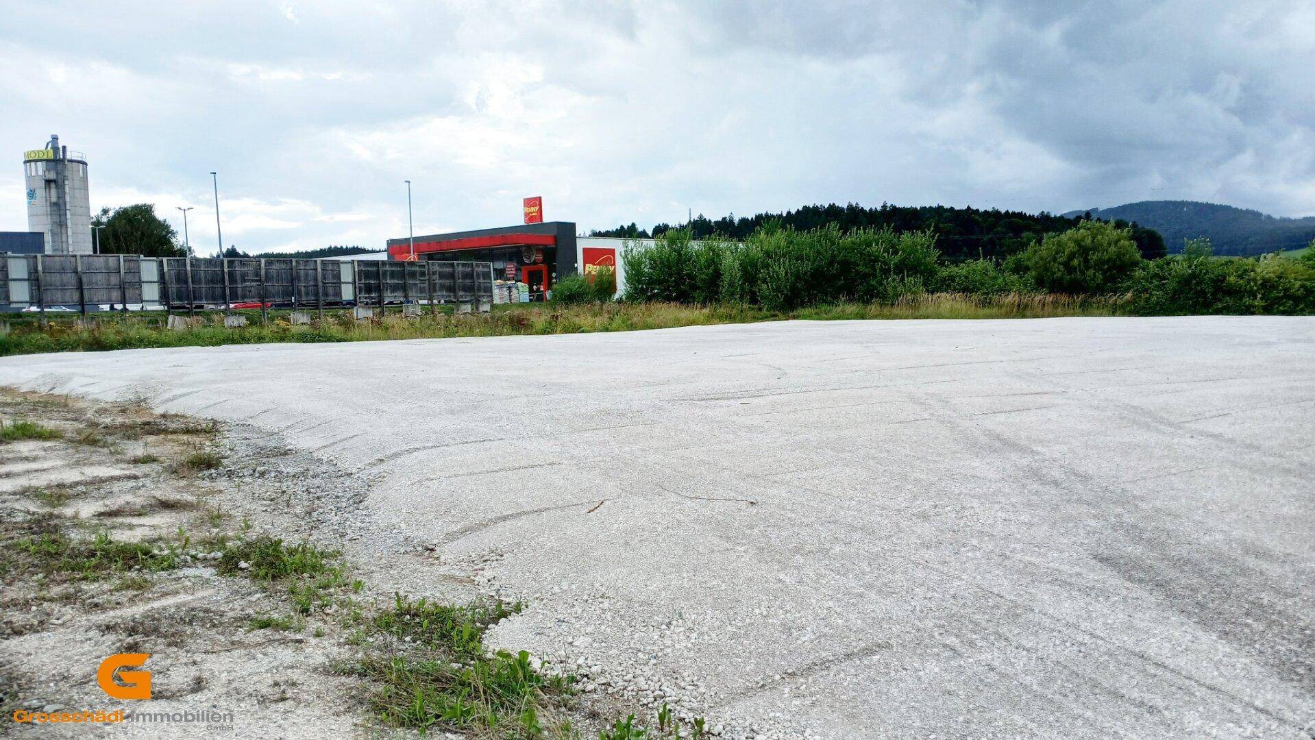 Leeres, gepflastertes Grundstück mit angrenzender Tankstelle und bewaldeter Hügellandschaft im Hintergrund.