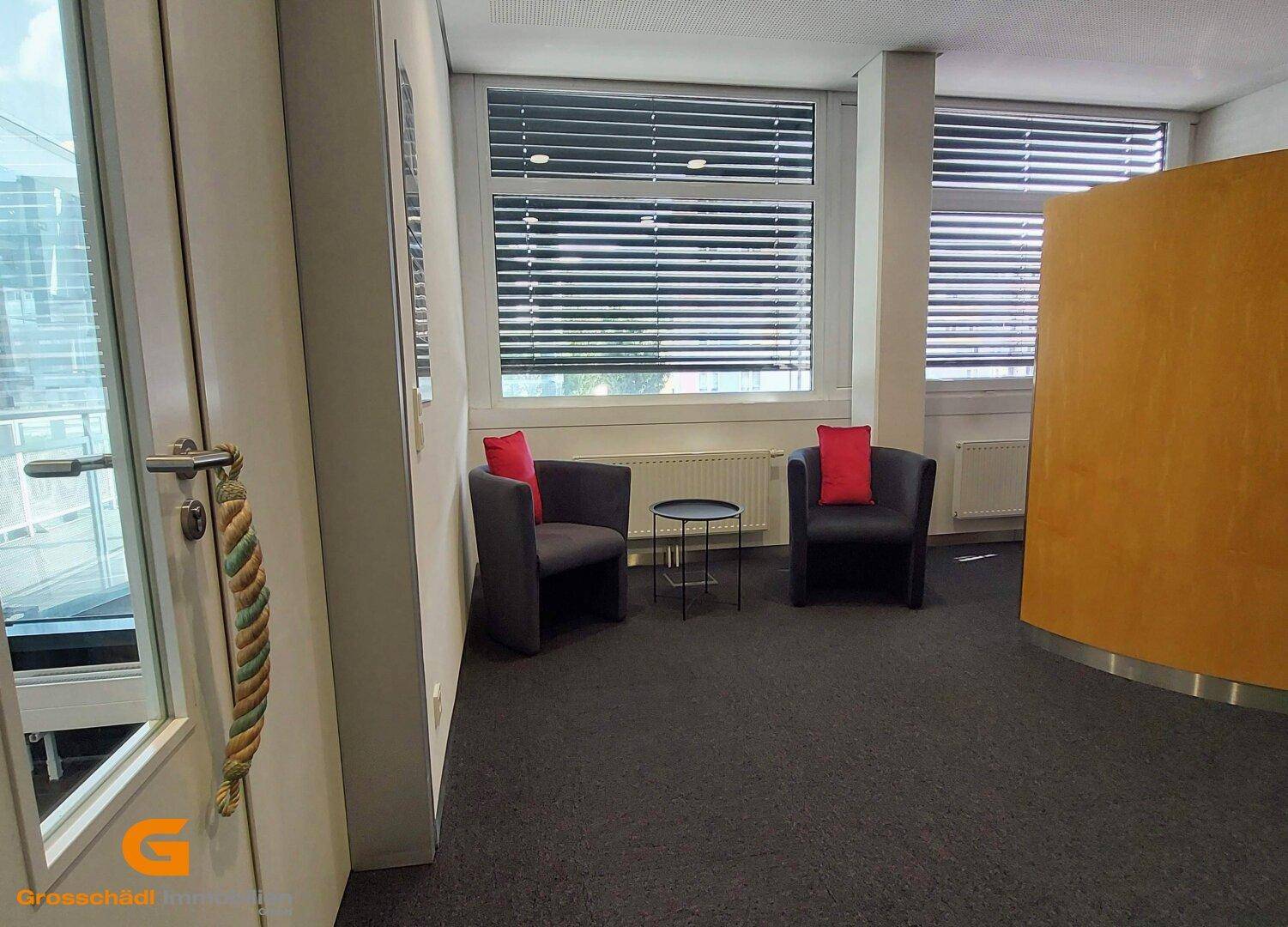 Gemütliche Sitzecke in einem Büro mit zwei grauen Sesseln, einem Beistelltisch und Fenstern mit Jalousien.