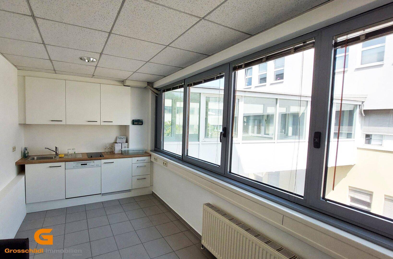 Kleine Einbauküche mit weißen Schränken, Holztheke und Fliesenboden, hell durch große Fenster mit Blick auf den Innenhof.