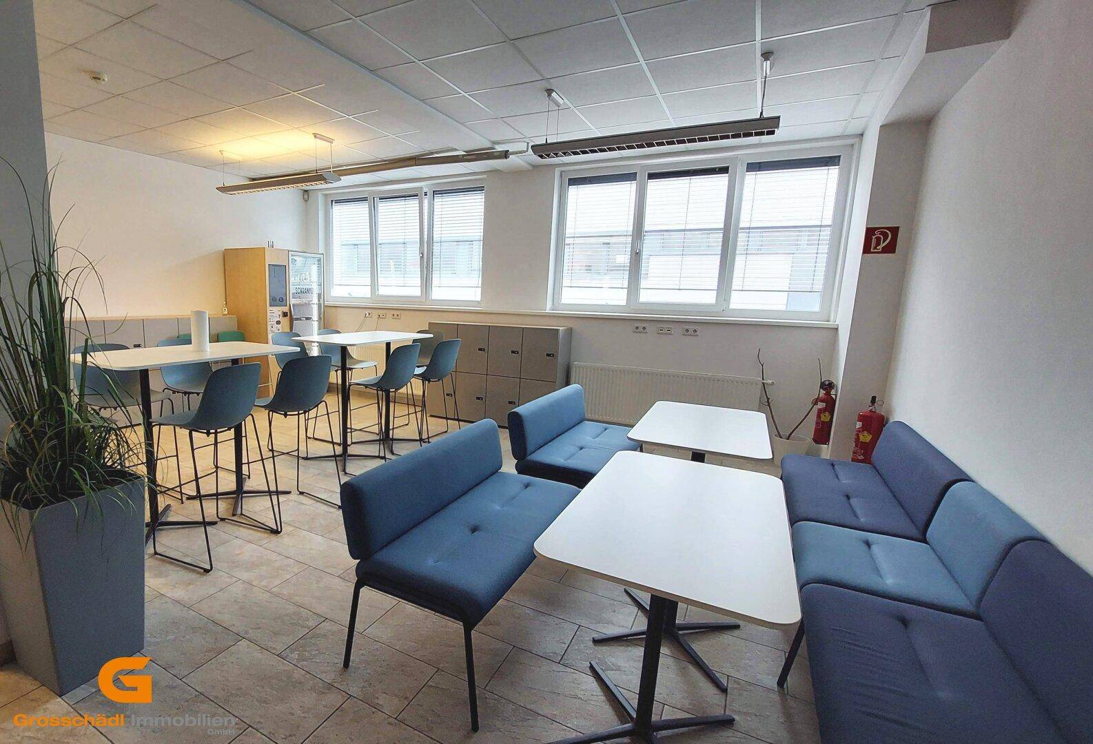 Heller Pausenraum mit verschiedenen Sitzgelegenheiten, Tischen und großen Fenstern.